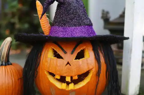 Pumpkin carved as a witch, with a pointed hat, yarn hair, and a carrot nose.