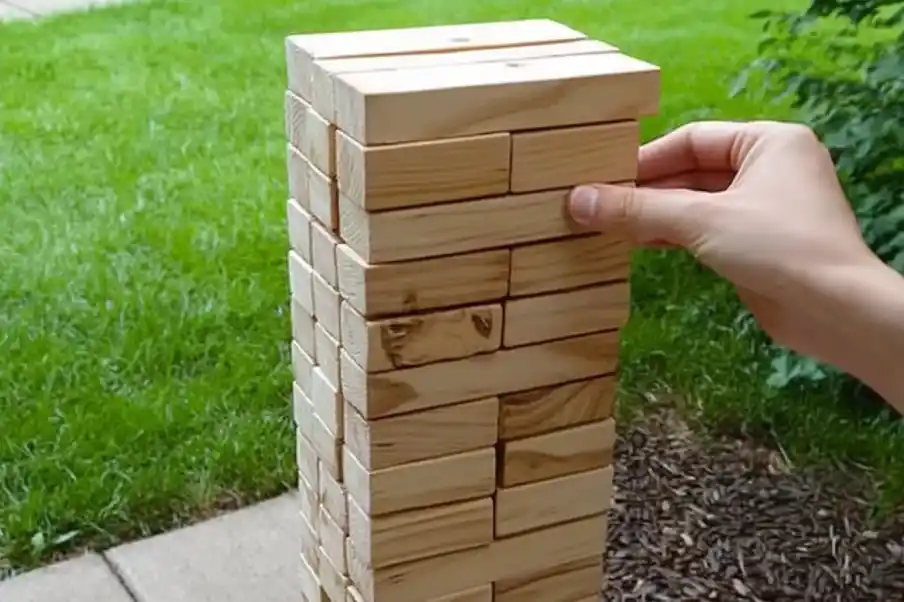 A hand carefully pulls a wooden block from a giant outdoor Jenga tower, set against a backdrop of lush green grass