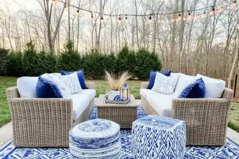 Elegant outdoor seating area with neutral-toned sofas, blue patterned rug, and poufs, framed by string lights against a natural backdrop