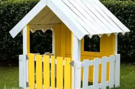 Handmade children's playhouse constructed from repurposed wooden pallets, painted in cheerful yellow and white, set up in a grassy backyard