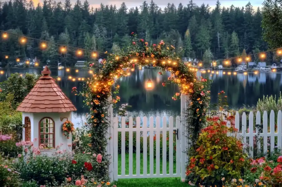 Idyllic garden with a white picket fence, archway adorned with greenery, and a lake view in the background.