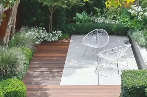 Small garden with wood and stone patios, white chairs, and lush greenery