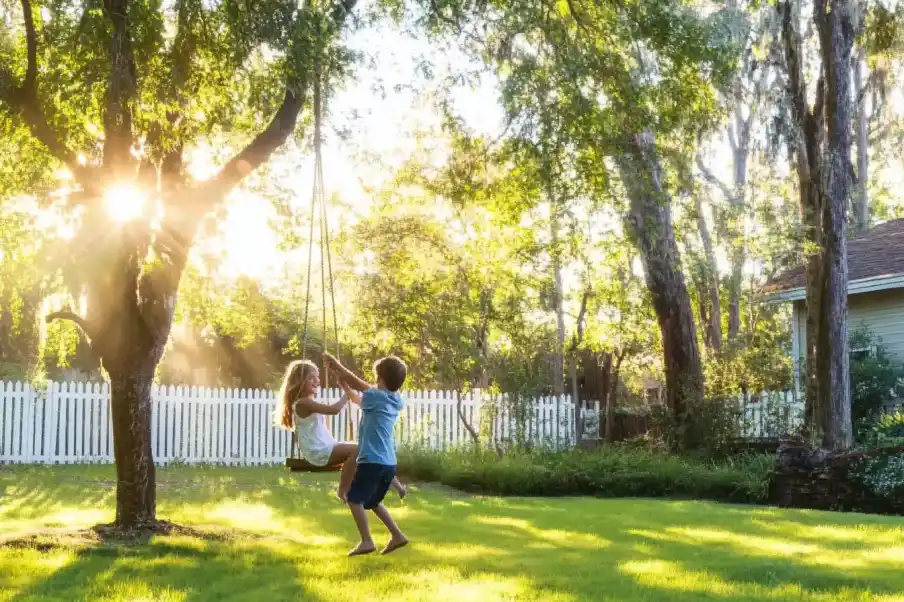 Sunlit backyard with children playing on a swing, surrounded by lush trees and a white picket fence, capturing a playful, carefree moment