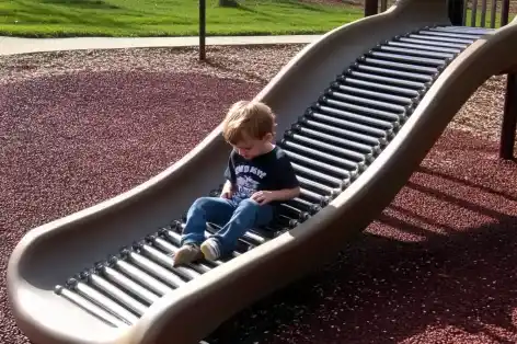 Young child sitting on a playground roller slide with metal rollers, enjoying a gentle ride down