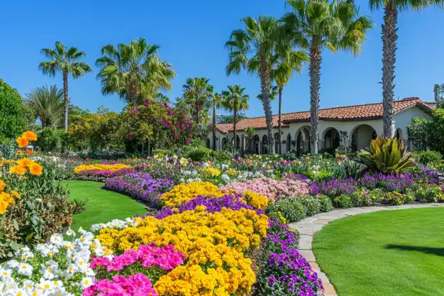 A stunning Mediterranean-style garden featuring vibrant flower beds filled with colorful blooms in shades of yellow, pink, purple, and white. The meticulously maintained lawn is bordered by a stone pathway that winds through the landscape. Tall palm trees frame the scene, with a charming villa in the background, showcasing its terracotta roof and elegant arches under a clear blue sky.