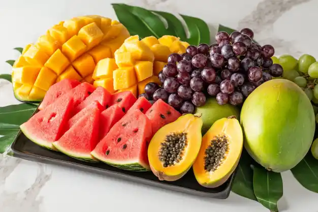 Vibrant tropical fruit platter featuring diced mangoes, sliced watermelon, papaya halves, grapes, and whole mango, displayed on banana leaves.