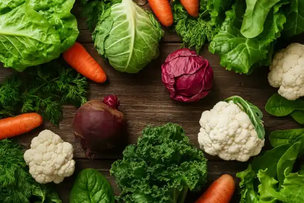 : A colorful assortment of fresh vegetables arranged on a wooden surface, including green lettuce, carrots, red cabbage, cauliflower, spinach, kale, and a beetroot.