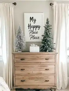 Wooden chest of drawers decorated with a frosted Christmas tree, a green evergreen tree, and a white gift box, with a framed sign reading "My Happy Place Place" on the wall above.