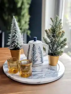 festive tabletop display with a marble tray featuring a teapot decorated with pine tree motifs, two small frosted Christmas trees in gold pots, and two amber glass cups.