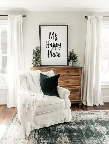  Minimalistic living room with a white armchair draped in a cozy throw, a black cushion, a wooden chest of drawers with small evergreen decor, and a framed sign reading "My Happy Place" above it.