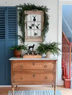 Rustic holiday decor featuring a wooden dresser topped with a white bowl of pinecones and a glass vase with evergreen sprigs, accompanied by a distressed blue shutter decorated with deer-themed art and greenery, situated in a cozy entryway with festive garlands on the staircase