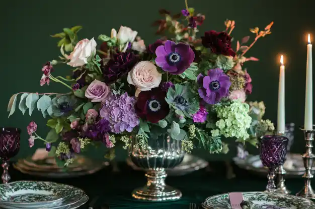 A lavish floral centerpiece featuring deep purple anemones, blush roses, hydrangeas, and greenery in a silver vase, elegantly displayed on a dining table set with vintage-style plates and candlesticks.