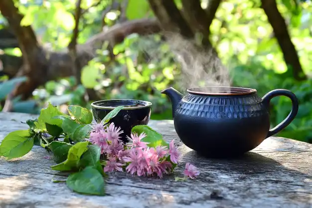 A black teapot with steam rising, accompanied by a small black cup, fresh herbs, and pink flowers, set on a rustic wooden table outdoors, surrounded by greenery.