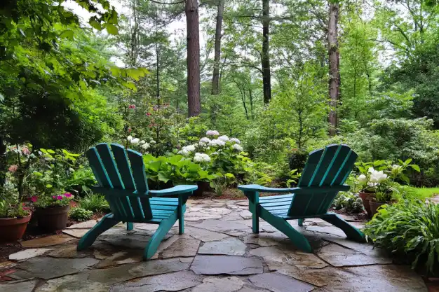 Two teal Adirondack chairs placed on a stone patio surrounded by a lush green garden with blooming white hydrangeas, colorful flowers, and tall trees creating a serene forest backdrop.