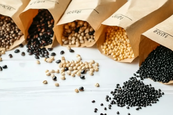 A collection of various seeds spilling out from labeled brown paper bags onto a white wooden surface. The seeds vary in size, shape, and color, including black, beige, and yellow varieties.