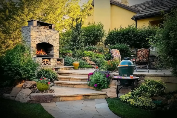 A beautifully landscaped backyard patio featuring a stone fireplace with a cozy fire burning. The space includes a tiered stone pathway leading up to an outdoor seating area with cushioned chairs, surrounded by lush greenery and colorful flowers. A green ceramic grill is placed nearby on a small table, suggesting a perfect spot for outdoor cooking and entertaining.