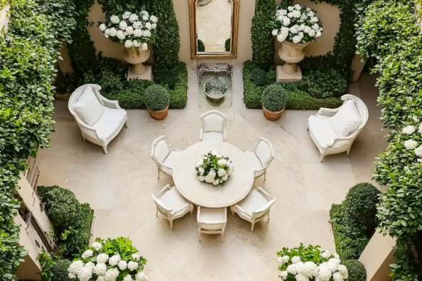 A beautifully designed courtyard garden featuring a circular white table surrounded by six chairs, two elegant white armchairs, and lush greenery.