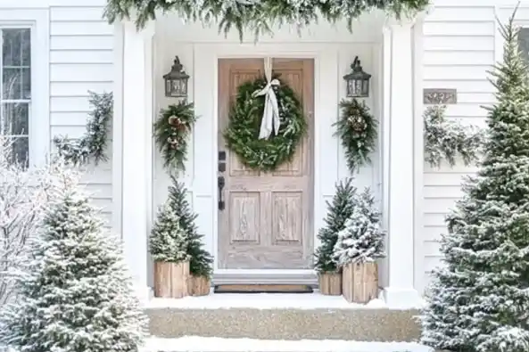 A snowy front porch decorated for Christmas with a wreath on the wooden door, garlands, and small evergreen trees in wooden planters.