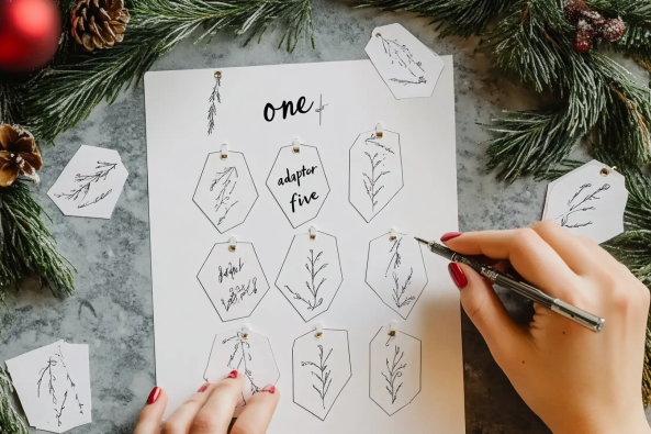 A person with red-painted nails is writing on a sheet of handmade gift tags featuring delicate botanical illustrations. The sheet is placed on a textured grey surface and surrounded by festive pine branches, pinecones, and red ornaments. Some tags are cut out and scattered around.