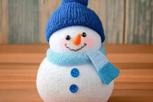 A cute handmade snowman crafted from a white sock, featuring a blue knitted hat, a matching blue scarf, and two blue buttons. The snowman has a cheerful smile with black eyes, rosy cheeks, and an orange nose resembling a carrot, sitting on a wooden surface with a rustic blue background.