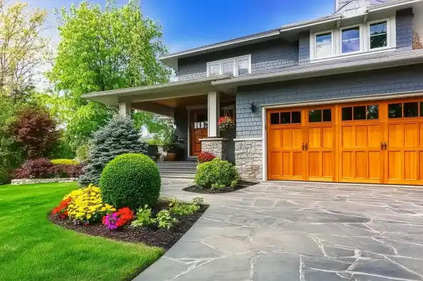 A beautifully landscaped front yard featuring a modern home with gray siding and wooden garage doors. The yard includes a manicured lawn, vibrant flower beds with colorful blooms, neatly trimmed shrubs, and a paved stone walkway leading to a welcoming porch adorned with potted plants. Title: Modern Home with Stunning Landscaping and Vibrant Flower Beds