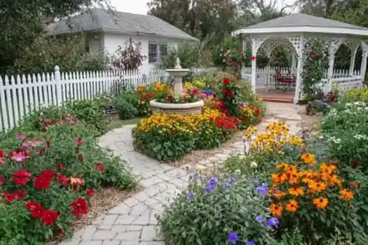 A charming cottage garden featuring colorful flower beds, a central fountain, a white picket fence, and a gazebo with a cozy seating area.