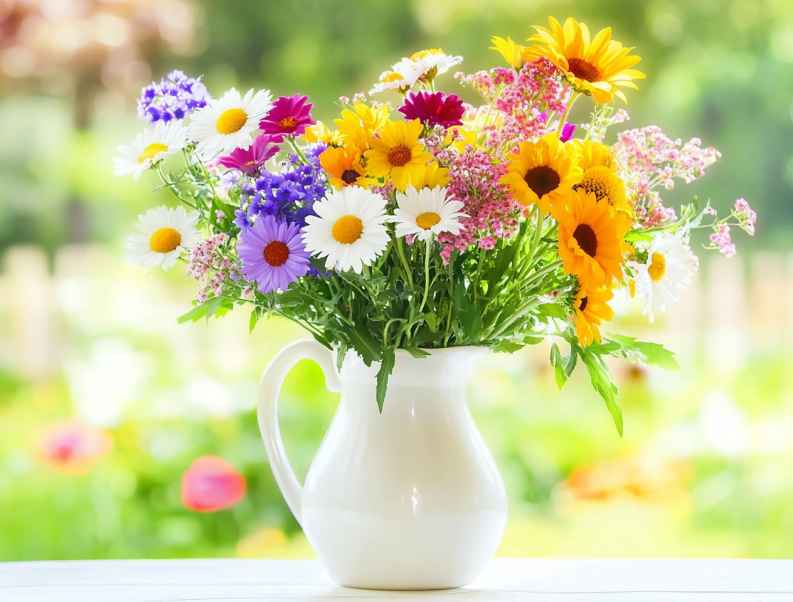 A cheerful bouquet of colorful wildflowers, including daisies, sunflowers, and other blooms, displayed in a simple white jug. The arrangement is set against a soft, sunny outdoor garden backdrop, adding a fresh and lively feel.