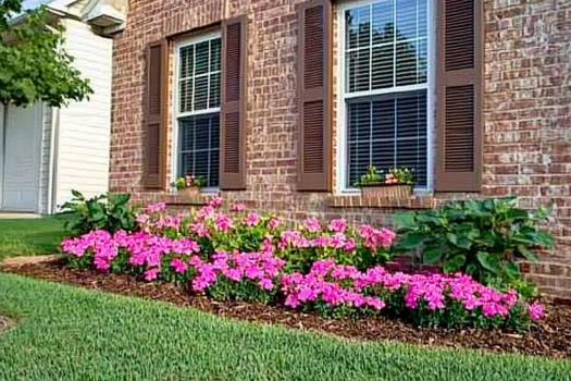 A neatly landscaped front yard featuring a brick home with brown shutters and large windows. A vibrant flower bed with bright pink blooms and green foliage lines the house, adding color and curb appeal. The lush green lawn enhances the welcoming atmosphere.