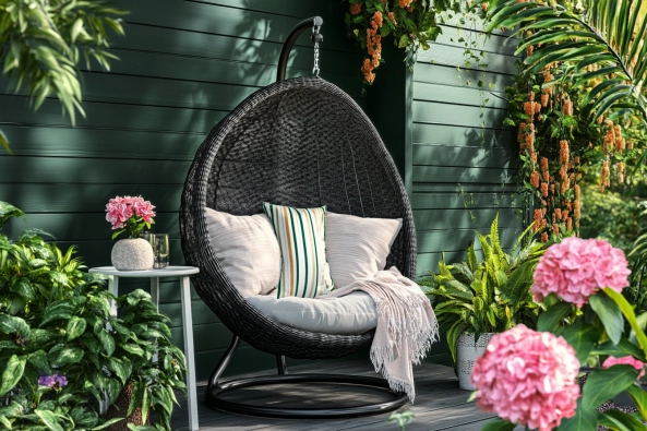 A beautifully designed outdoor nook featuring a black wicker hanging egg chair with plush cushions and a soft throw blanket. Surrounded by lush greenery and blooming pink hydrangeas, the setting exudes a tranquil and inviting atmosphere. A small round side table with a decorative plant and stone adds a stylish touch. The dark green wooden backdrop enhances the cozy ambiance, making it a perfect relaxation spot.