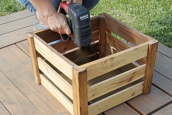 A person using a power sander to smooth the surface of a handmade wooden crate. The crate, made of light and dark wooden slats, is placed on a wooden deck with a grassy lawn in the background. The person is wearing jeans and is carefully working on refining the wood, possibly for a finishing coat or stain. The setting suggests an outdoor DIY woodworking project.