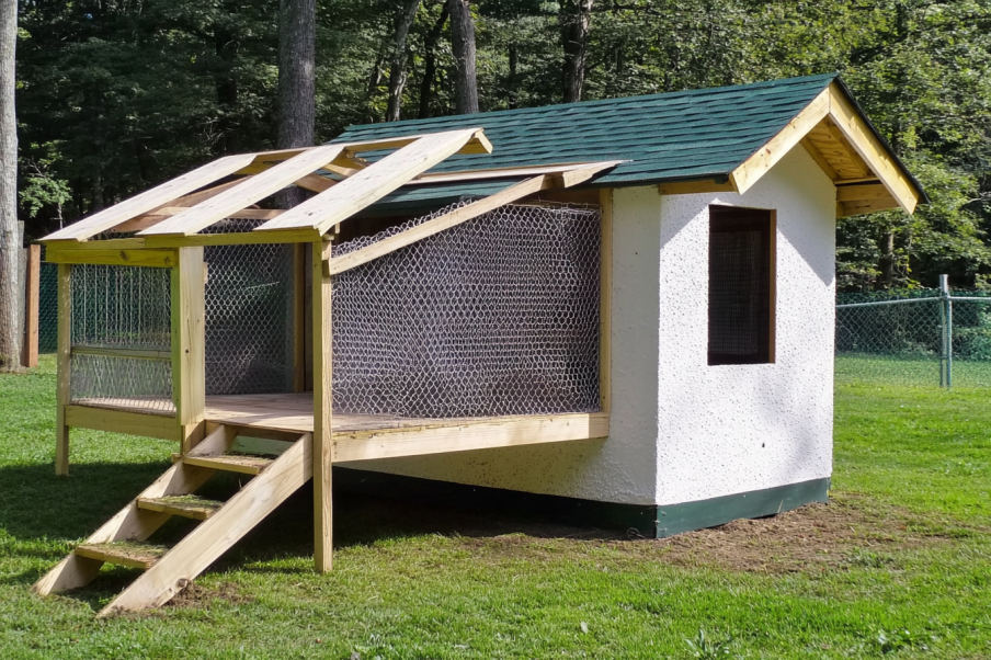 A well-built elevated chicken coop featuring a white stucco exterior, green shingled roof, and a secure wire mesh enclosure. A wooden staircase leads up to a raised platform with an attached run, partially covered by an unfinished wooden roof frame. The coop is set in a grassy backyard surrounded by trees and fencing, providing a spacious and natural environment for poultry.