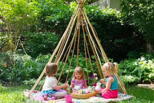 Three young girls enjoying a tea party in a lush green backyard, sitting on a colorful blanket under a handmade bamboo teepee. The teepee structure is made from wooden sticks, creating a cozy play space. The children are engaged in imaginative play, surrounded by nature and dappled sunlight filtering through the trees. A picnic basket, toy teapot, and cups complete the charming outdoor setting.